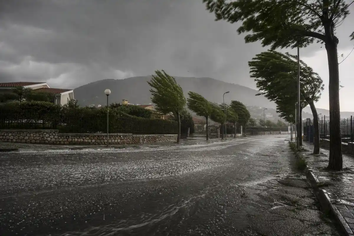 Strada urbana sotto un temporale con grandine, alberi piegati dal vento e nuvole scure sulle colline