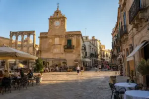Piazza in pietra al tramonto con edificio storico con orologio, tavolini all’aperto, passanti e colonne antiche