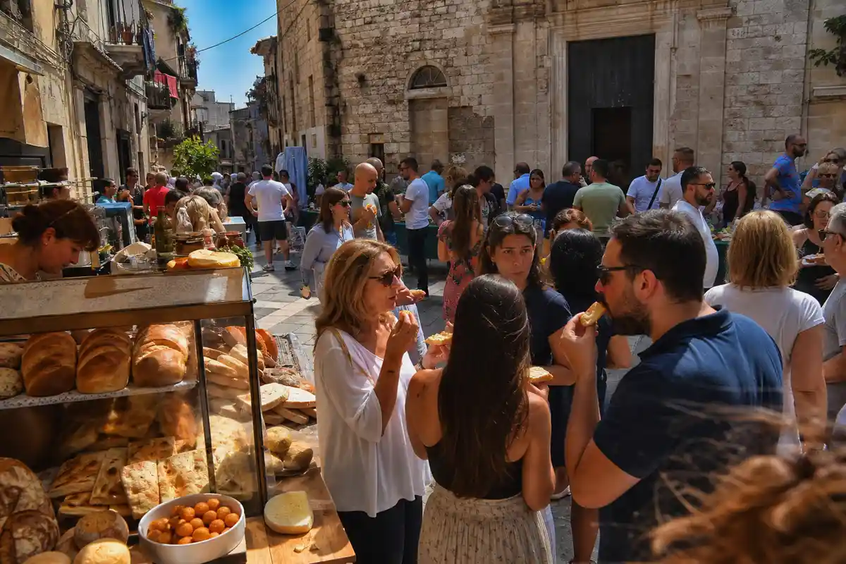 Gente che mangia pane e focaccia in una piazza di pietra con banco di forno e vicoli sullo sfondo