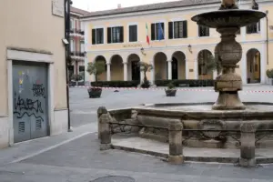 Piazza con fontana e municipio sullo sfondo, porta tecnica con graffiti e nastro bianco e rosso