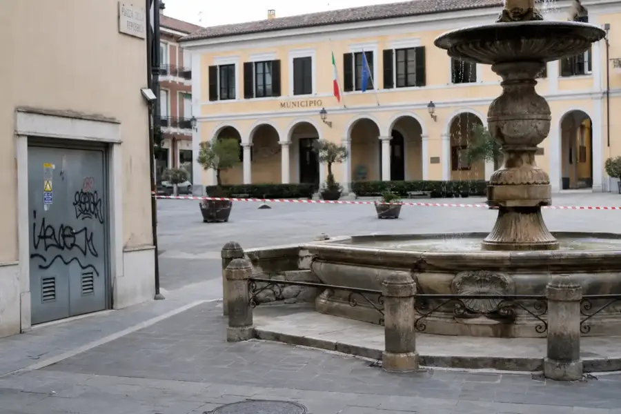 Piazza con fontana e municipio sullo sfondo, porta tecnica con graffiti e nastro bianco e rosso