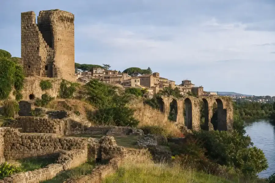 Ruderi in pietra con torre, archi di un antico acquedotto e fiume accanto a un borgo su una collina