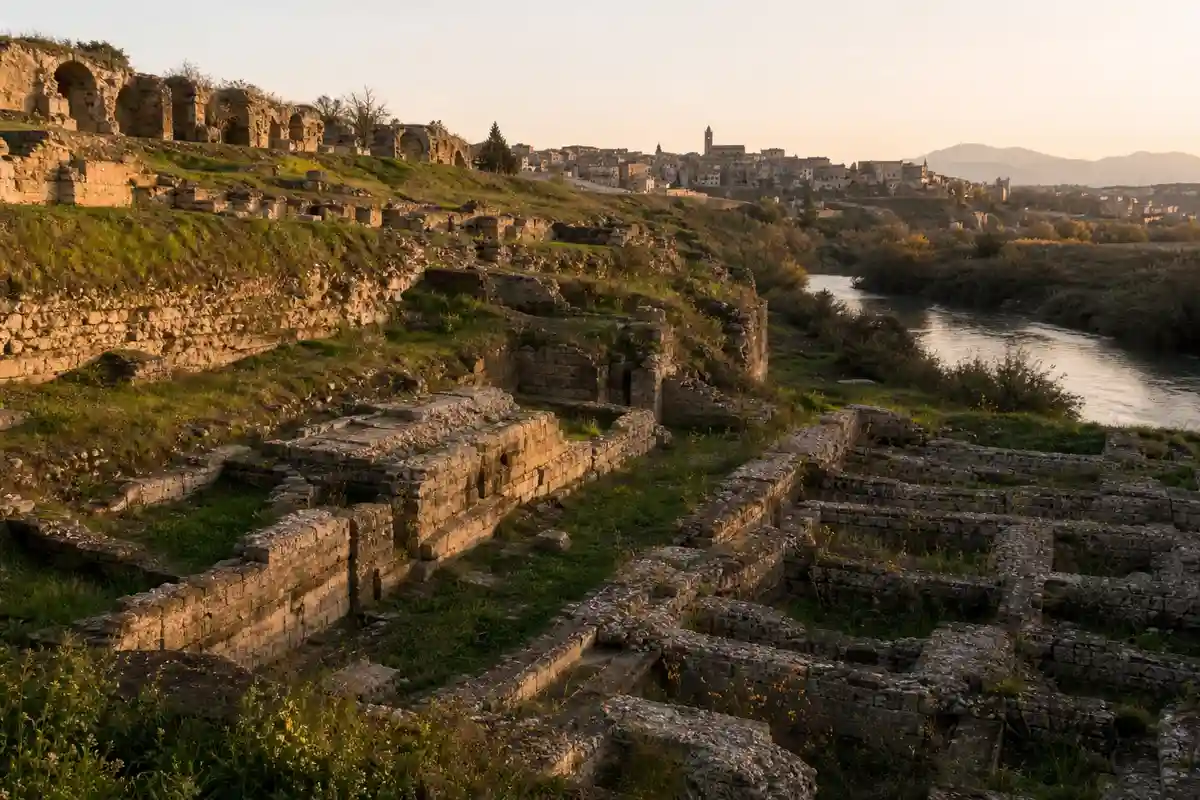 Rovine in pietra su un pendio erboso vicino a un fiume, con un borgo in lontananza al tramonto