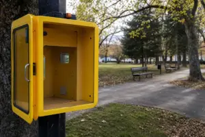 Techetta gialla per defibrillatore vuota e aperta su un palo, con panchine e vialetto in un parco