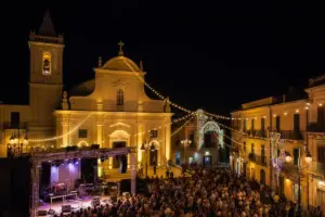 Piazza di sera con chiesa illuminata, luci festive, palco per concerto e folla riunita