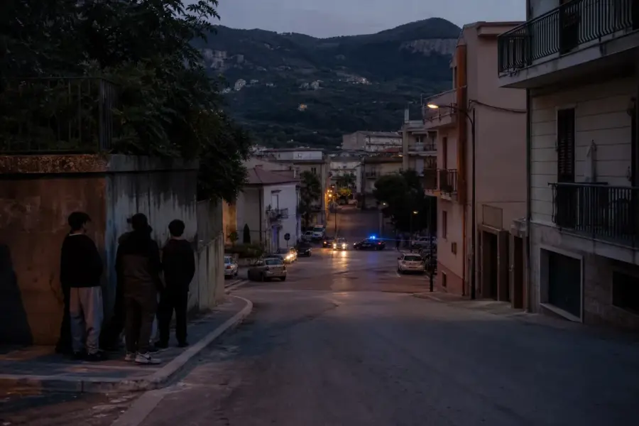 Strada di un paese al crepuscolo con tre ragazzi in ombra sul marciapiede e lampeggianti blu in lontananza