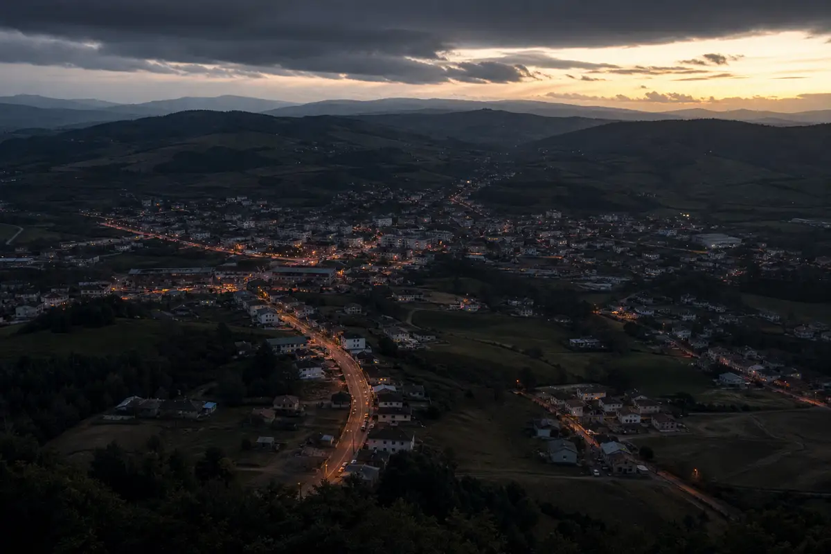 Veduta aerea al tramonto di una cittadina tra colline, con strade illuminate e cielo scuro