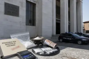 Buste di polvere bianca, bilancino e fascicolo “Processo per spaccio di cocaina” davanti a un tribunale con auto della