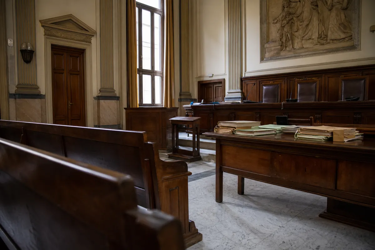 Aula di tribunale vuota con banchi in legno, tavolo dei giudici e fascicoli, illuminata da una grande finestra