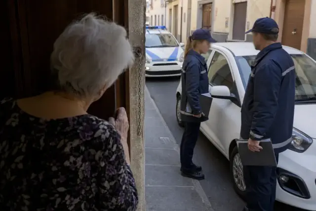 Anziana guarda da una porta socchiusa due persone in divisa vicino a un’auto bianca, con pattuglia sullo sfondo