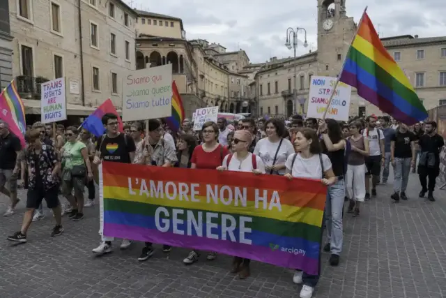 Corteo Pride in una piazza con edifici storici, bandiere arcobaleno e striscione “L’amore non ha genere