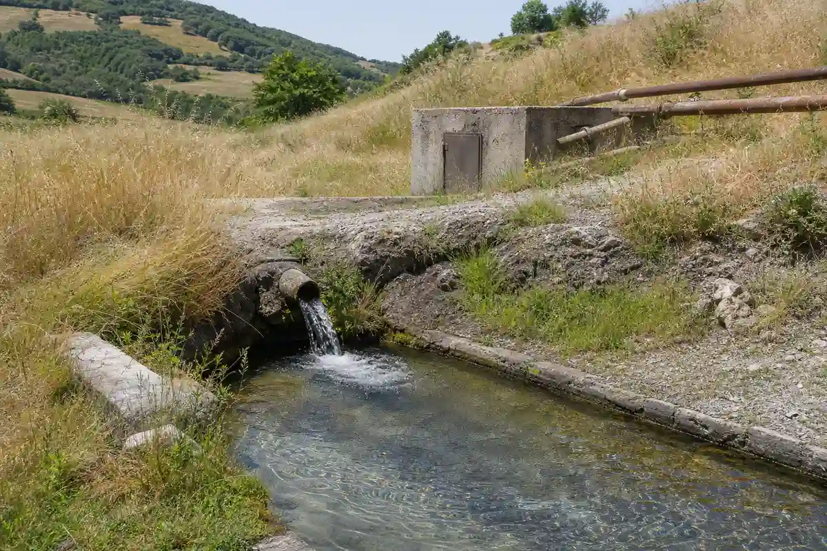 Acqua di sorgente che esce da un tubo in un canale di pietra, tra colline e tubazioni in un prato estivo
