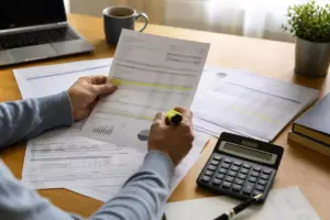 Mani evidenziano voci su documenti finanziari su una scrivania con calcolatrice, laptop e tazza di caffè