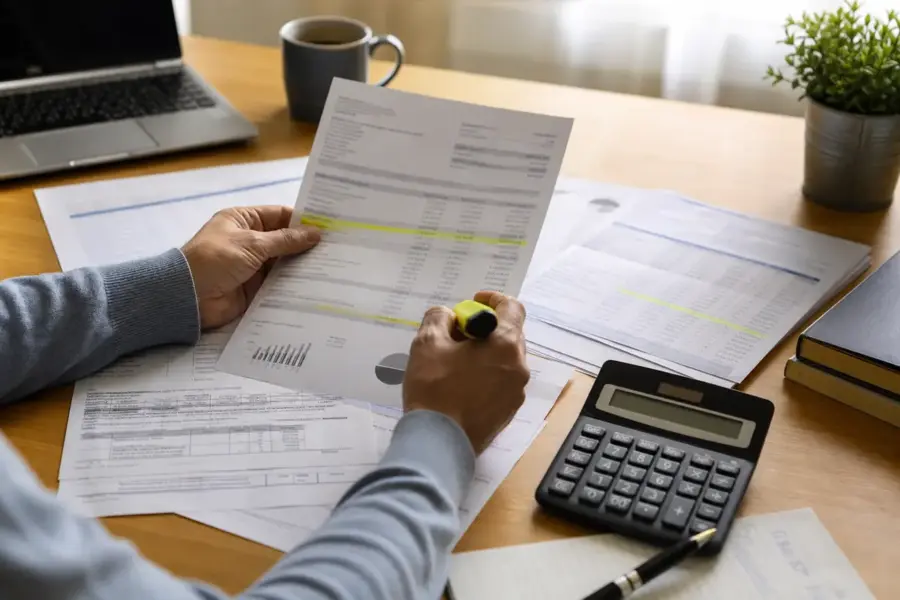 Mani evidenziano voci su documenti finanziari su una scrivania con calcolatrice, laptop e tazza di caffè