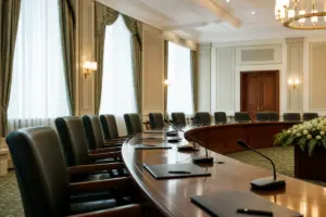 Sala riunioni istituzionale con tavolo curvo, microfoni, cartelline e sedie vuote vicino a grandi finestre