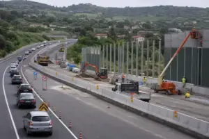 Cantiere autostradale con operai e macchinari per barriere antirumore, traffico deviato tra coni e corsie ridotte
