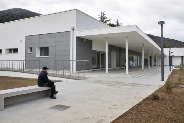Anziano seduto fuori da un centro sanitario moderno quasi vuoto, con due operatori in camice all’ingresso