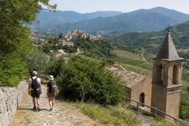 Escursionisti su sentiero in pietra verso un borgo collinare, con campanile e montagne verdi sullo sfondo