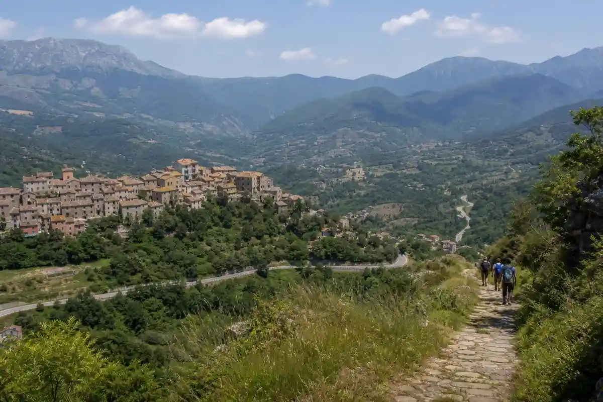 Sentiero panoramico con escursionisti e borgo collinare tra montagne verdi e strade tortuose