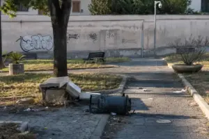 Panchina rotta e cestino divelto su un vialetto di parco, con graffiti sul muro e una telecamera sullo sfondo