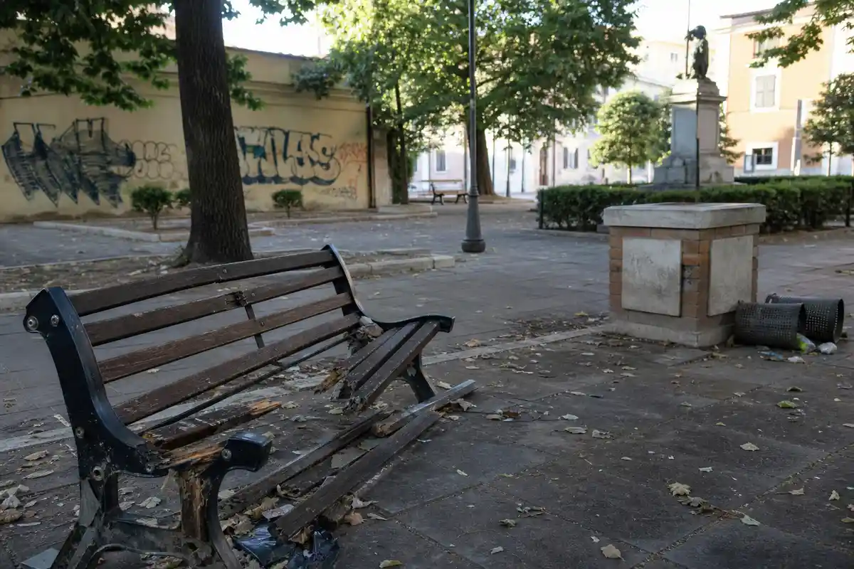 Panchina rotta in un parco cittadino, con foglie e rifiuti a terra e muro con graffiti sullo sfondo