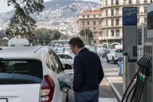 Tassista fa rifornimento a un taxi bianco in una stazione di servizio, con traffico cittadino sullo sfondo