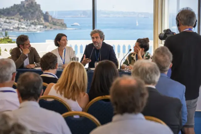 Relatore al microfono in un panel sul cinema con pubblico in sala e vista sul mare da una terrazza