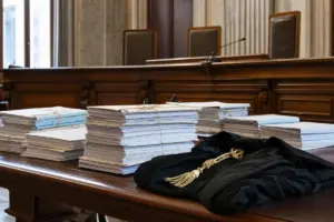 Fascicoli legali impilati su un tavolo in aula di tribunale, con toga nera in primo piano e banco dei giudici sullo sfondo