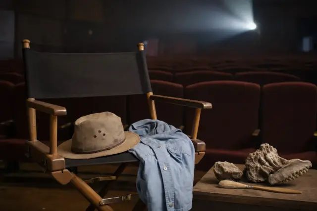 Sedia da regista vuota in un cinema, con cappello beige e camicia azzurra, accanto a fossili e pennello
