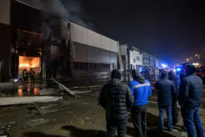 Magazzino industriale in fiamme di notte con vigili del fuoco, fumo denso e persone che osservano a distanza