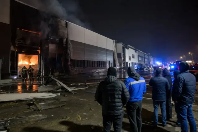 Magazzino industriale in fiamme di notte con vigili del fuoco, fumo denso e persone che osservano a distanza