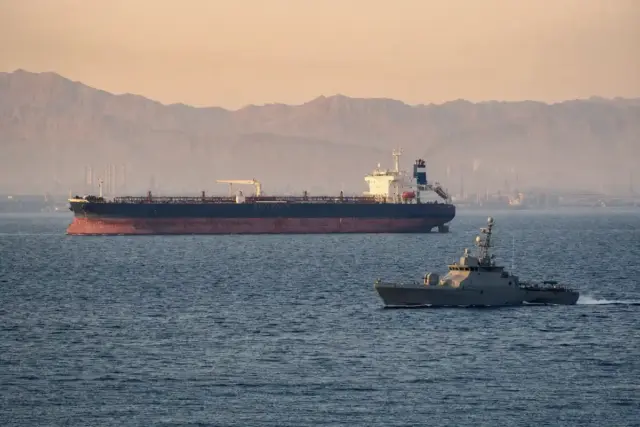 Petroliera in mare con una piccola nave militare di pattuglia in primo piano e costa montuosa nella foschia