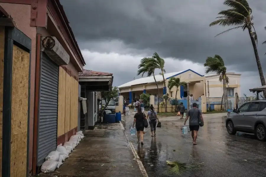 Residenti con bottiglie d’acqua camminano verso una scuola mentre palme piegate e cielo scuro annunciano il tifone