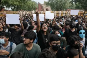 Giovani manifestanti a Delhi con cartelli bianchi, alcuni con maschere, davanti a transenne e poliziotti