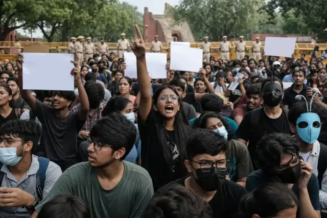 Giovani manifestanti a Delhi con cartelli bianchi, alcuni con maschere, davanti a transenne e poliziotti