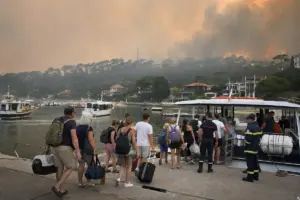Evacuati con valigie e zaini salgono su un traghetto in un piccolo porto, con fumo d’incendio sullo sfondo