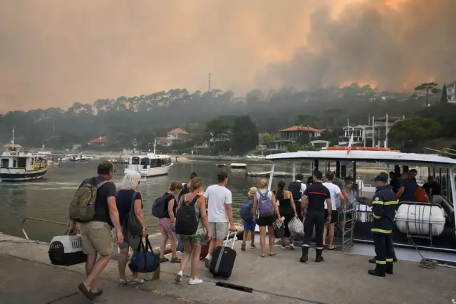 Evacuati con valigie e zaini salgono su un traghetto in un piccolo porto, con fumo d’incendio sullo sfondo