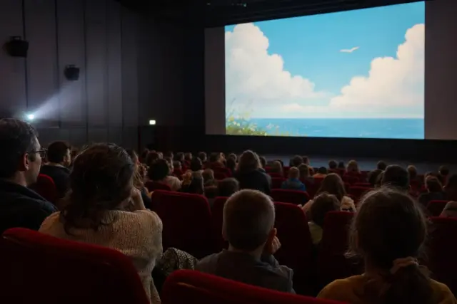 Bambini e genitori in sala cinema guardano un film: sullo schermo cielo e mare animati con un gabbiano in volo