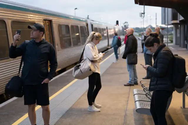 Pendolari su una banchina con treno fermo, controllano gli smartphone all’alba durante un disservizio di rete
