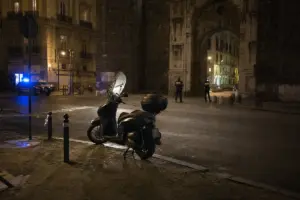 Scooter parcheggiato di notte davanti a un grande arco in pietra, con due poliziotti e lampeggianti blu sullo sfondo
