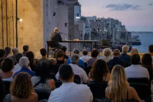 Pubblico ascolta un relatore su un palco all’aperto al tramonto, con microfono, libri e mare sullo sfondo