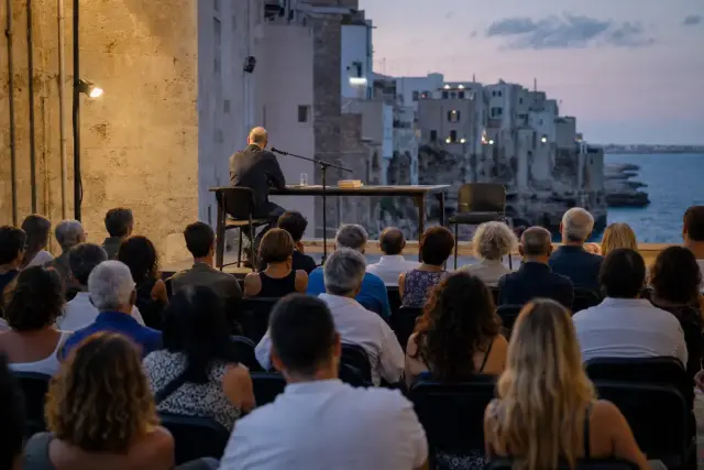 Pubblico ascolta un relatore su un palco all’aperto al tramonto, con microfono, libri e mare sullo sfondo