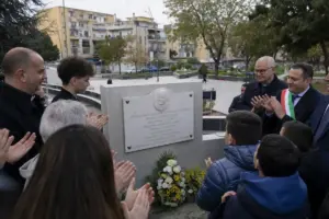 Persone in piazza applaudono davanti a una targa commemorativa su un muretto, con mazzo di fiori ai piedi