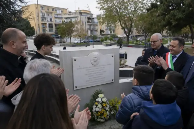 Persone in piazza applaudono davanti a una targa commemorativa su un muretto, con mazzo di fiori ai piedi