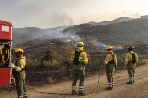 Vigili del fuoco su una strada sterrata osservano colline bruciate con fumo e case isolate sullo sfondo