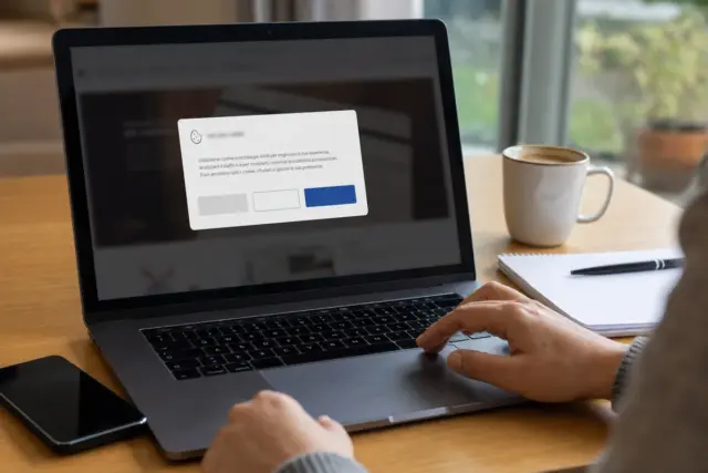 Mano sul trackpad davanti a un laptop con pop-up di consenso ai cookie su una scrivania con tazza e quaderno
