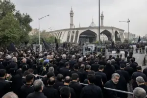 Folla in abiti neri con bandiere nere e transenne davanti al Grand Mosalla di Teheran, sotto cielo grigio