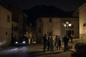 Residenti fuori in una piazzetta di paese di montagna di notte, con auto a fari accesi e lampioni