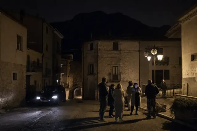 Residenti fuori in una piazzetta di paese di montagna di notte, con auto a fari accesi e lampioni