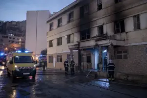 Vigili del fuoco e ambulanze davanti a un edificio annerito dal fumo dopo un incendio, con strada bagnata all’alba